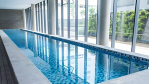 A long indoor swimming pool with clear blue water, surrounded by large windows and a wooden deck. - Luxury studio apartment in Vila Olímpia - panoramic view and garage. (São Paulo)