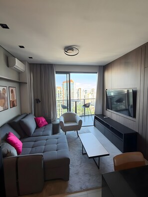 A modern living room with a grey sofa, a small armchair, a TV, and a balcony with a city view. - Luxury studio apartment in Vila Olímpia - panoramic view and garage. (São Paulo)