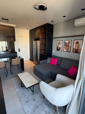 A modern living room with a grey sofa, white armchair, and a coffee table. There is a kitchen area with a refrigerator and cabinets. - Luxury studio apartment in Vila Olímpia - panoramic view and garage. (São Paulo)