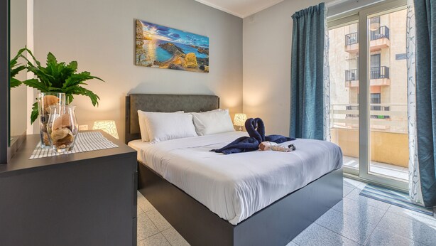 Quarto de hotel com uma cama, um quadro na parede, uma planta num apoio lateral e vista pela janela.