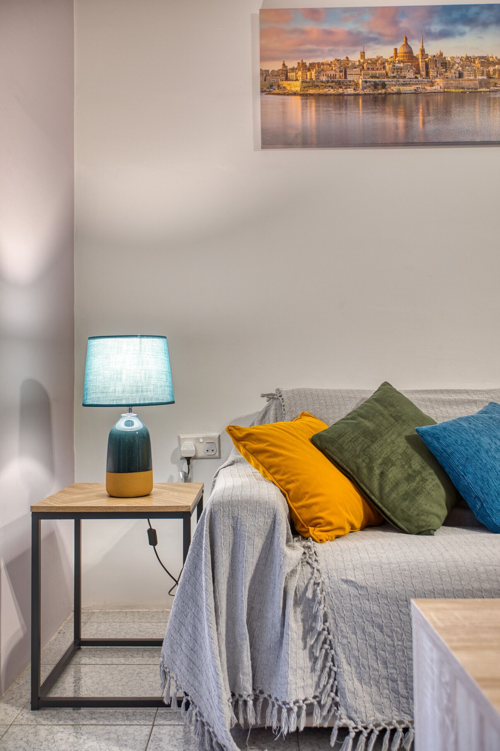 A neatly arranged bedroom with a bed, a bedside table with a lamp, a power outlet, and a cityscape painting on the wall.