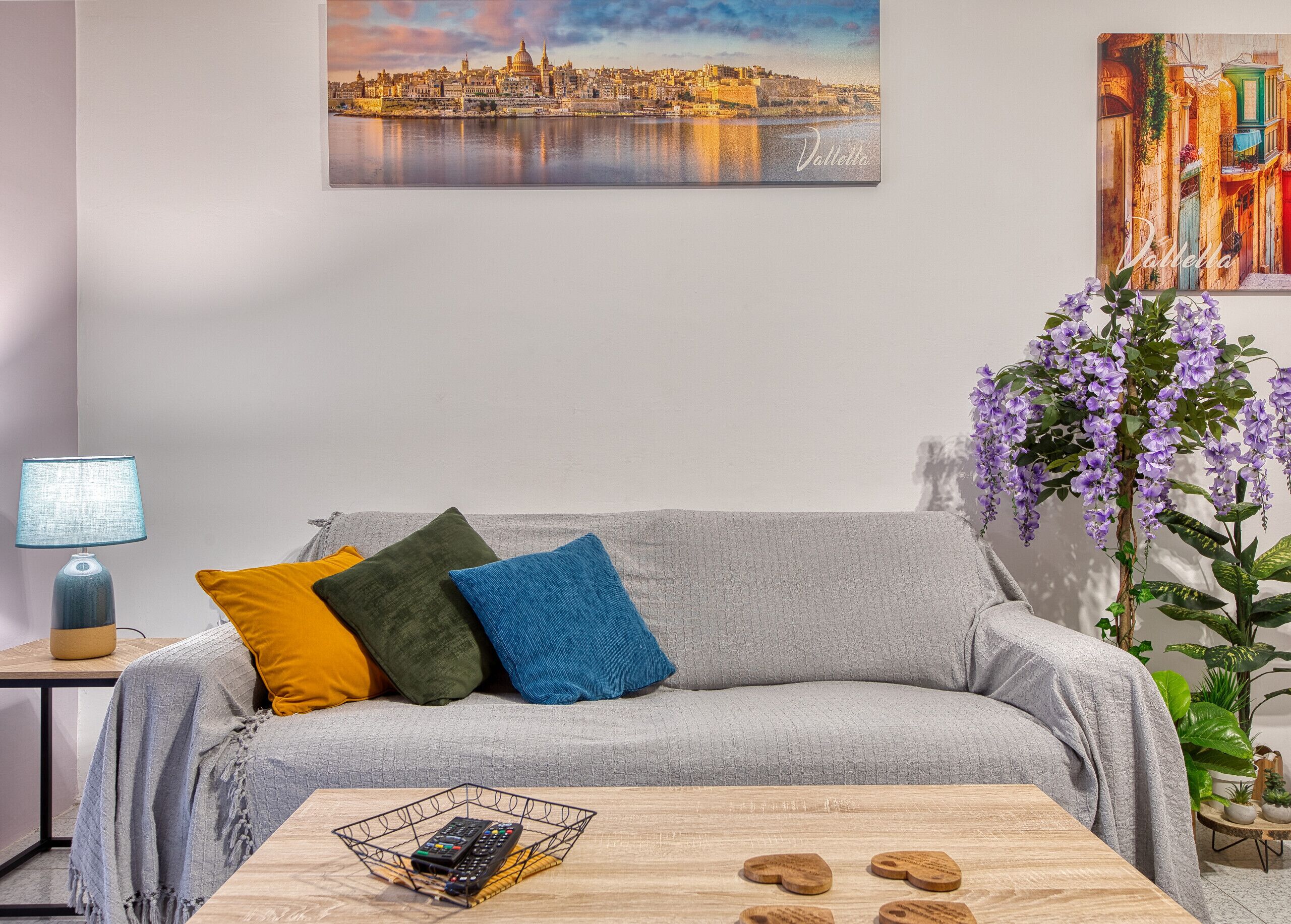 A living room with a grey sofa, colorful cushions, a coffee table with remote controls, a lamp, and a cityscape painting.