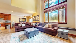 A spacious living room with a sectional sofa, a coffee table, and a large window with a forest view. - Golf Course Retreat, Private Hot Tub, BBQ, Views, Garage, Pet Friendly (Mammoth Lakes)
