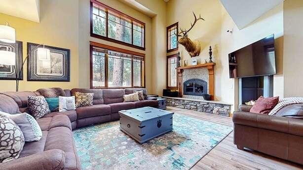 A spacious living room with a fireplace, large windows, and a mounted deer head. - Golf Course Retreat, Private Hot Tub, BBQ, Views, Garage, Pet Friendly (Mammoth Lakes)