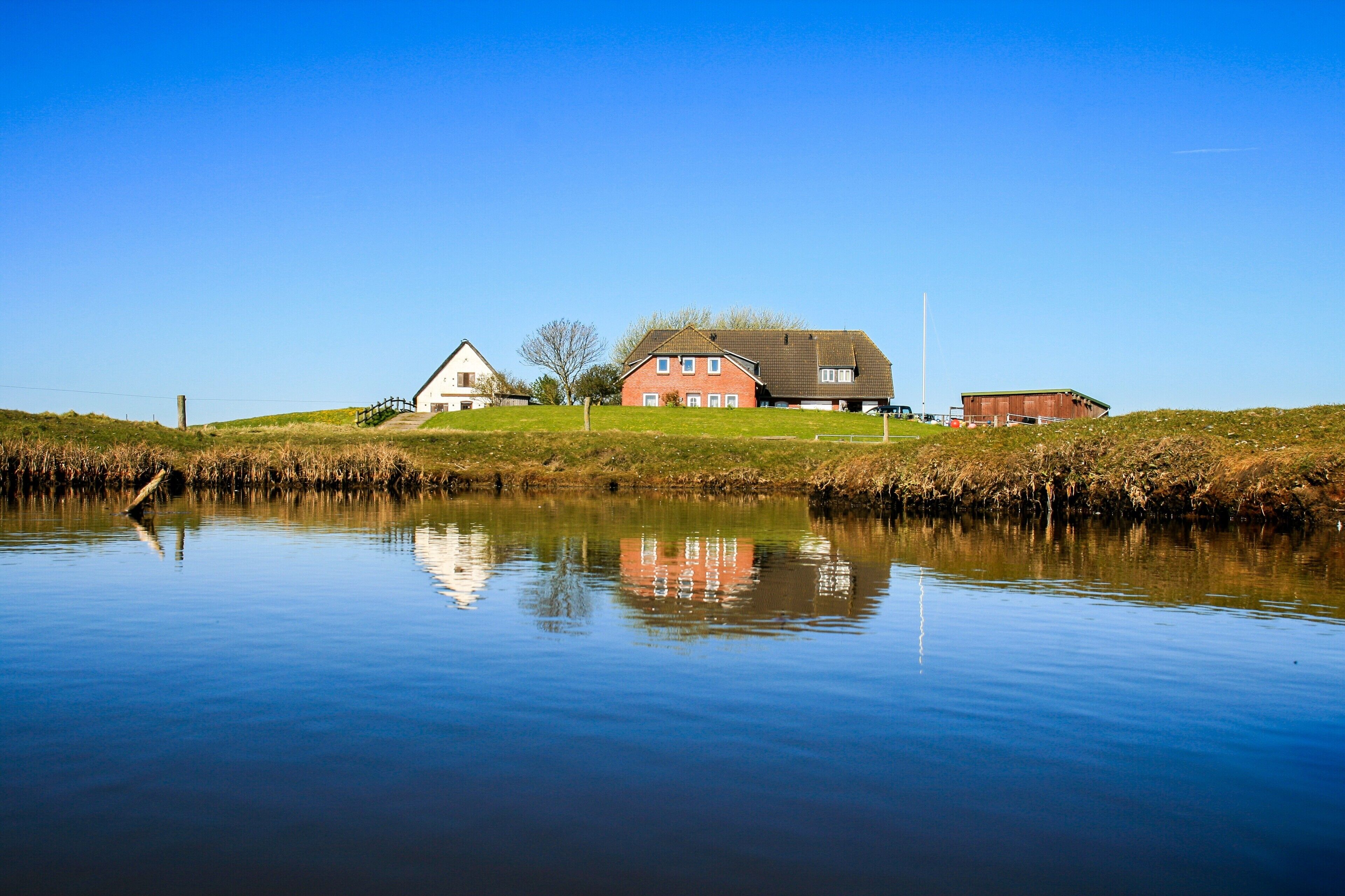 A calm lake with a reflection of buildings, including a red house and a white house, surrounded by grassy banks.
