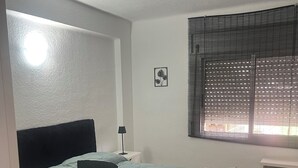 Ein Schlafzimmer mit einem Bett, einem Nachttisch, einer Lampe, einem Fenster mit Jalousien und einem kleinen Bild an der Wand.