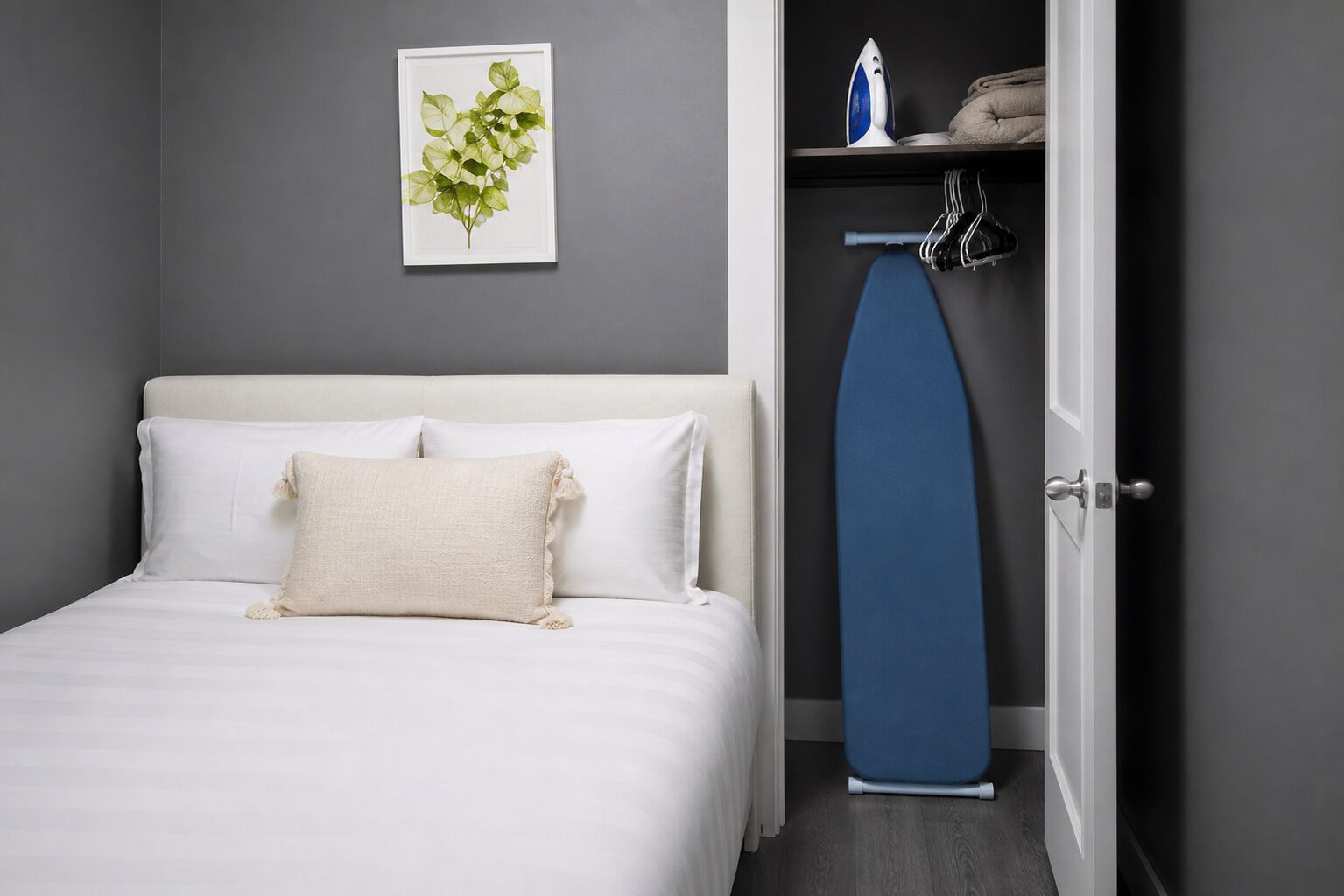 Camera da letto con un letto, cuscini, un quadro appeso al muro, una porta con un asse da stiro blu e uno scaffale con un ferro da stiro e asciugamani.