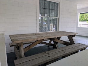 A wooden picnic table with benches in a covered porch area with white siding walls and a window with patterned curtains. - Grand Village Home. Central Location. EV Charger. (Corinth)