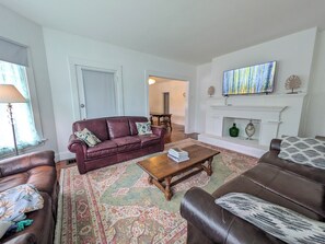 A living room with a fireplace, a television, and a dining area in the background. - Grand Village Home. Central Location. EV Charger. (Corinth)