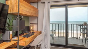 Ein Zimmer mit Schreibtisch aus Holz, Computermonitoren, Tastatur, einer Pflanze und Meerblick durch eine Schiebetür.
