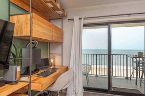 A room with a wooden desk, computer monitors, a keyboard, a plant, and a view of the ocean through a sliding glass door. - Oceanfront Condo with Indoor Pool (Ocean City)