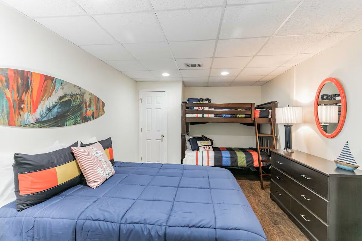 A bedroom with bunk beds, a bed with a blue quilt, a dresser, a mirror, and a colorful painting on the wall.