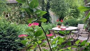 Een tuinpatios met een ronde tafel en stoelen, omgeven door weelderig groen en bloeiende planten.
