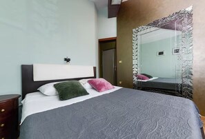 Un llit ben fet amb un capçal, dos almoinos, una taula de nit i un mirall gran amb un marc decoratiu.