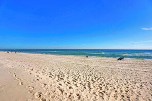 Ein weiter, sandiger Strand mit Fußabdrücken, ruhigem Wasser und blauem, wolkenlosem Himmel.