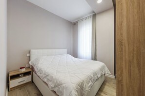 A bedroom with a bed, a bedside table, a nightstand, a drawer, and a window with curtains. - Cozy Wave Retreat | Beach 2 Min Walk by PikHost (Qerret)