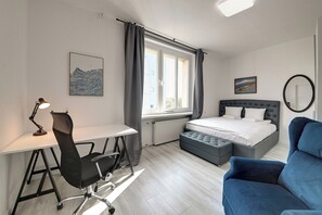 A bedroom with a bed, a desk, a chair, a blue armchair, a window with curtains, and a wall-mounted mirror. - RentPlanet - Apartament przy pl Legionów (Wrocław)