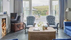 A living room with a fireplace, two armchairs, a sofa, a television, and a view of the countryside through large windows. - Luxury Coastal Cottage With Harbour Views (St Austell)