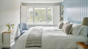 A bedroom with a large bed, a window with shutters, a chair, a bedside table, and a lamp. - Luxury Coastal Cottage With Harbour Views (St Austell)