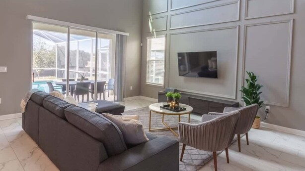 A living room with a gray sofa, a TV mounted on the wall, and a dining area with a table and chairs. - Luxury 5BR Private Pool Disney Area (Kissimmee)