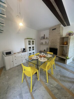 A dining area with a glass table and yellow chairs, a white cabinet with glass doors, a brick fireplace, and a white ceiling with exposed beams. - Dwelling Forest on the Blue (Ameglia)
