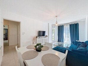 Un salon moderne avec une table à manger blanche, un canapé bleu et un téléviseur posé sur un meuble blanc.
