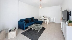 En moderne stue med en blå sofa, et glasbord, et spiseområde med et hvidt bord og stole, samt et køkkenområde med komfur og fjernsyn.