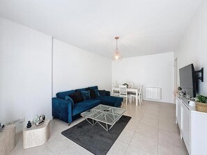 Un salon moderne avec un canapĂ© bleu, une table basse en verre, un coin repas avec une table et des chaises blanches, et un coin cuisine Ă©quipĂ© dâune cuisiniĂšre et dâun tĂ©lĂ©viseur.