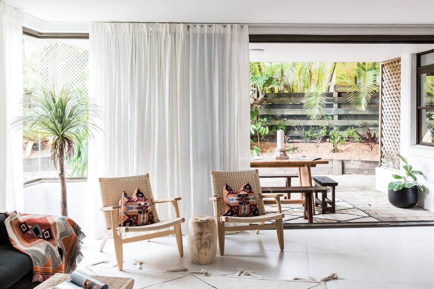 A room with a large window, two wicker chairs with patterned cushions, a wooden table, and a potted plant.