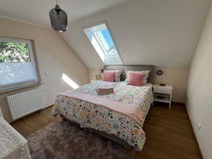 Ein Schlafzimmer mit schrägen Wänden, einem Bett mit floralem Bettbezug, einem Nachttisch und einem Fenster mit Jalousien.