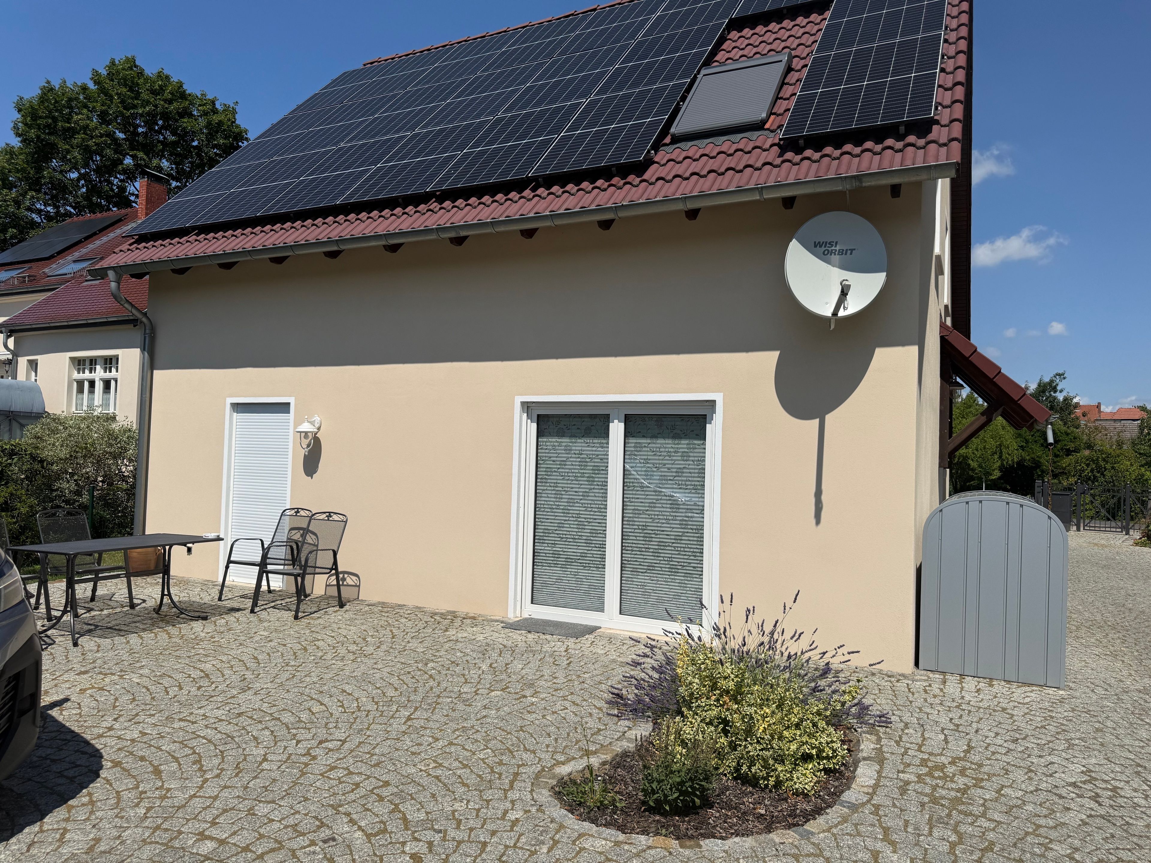 Une maison avec des panneaux solaires sur le toit, une antenne parabolique et une terrasse dallée aménagée avec du mobilier d’extérieur.