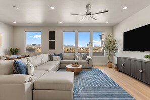 A modern living room with a sectional sofa, a flat-screen TV, a ceiling fan, and large windows offering a city view. - Vacation Mode: Private Golf Cart + Hot Tub (St. George)
