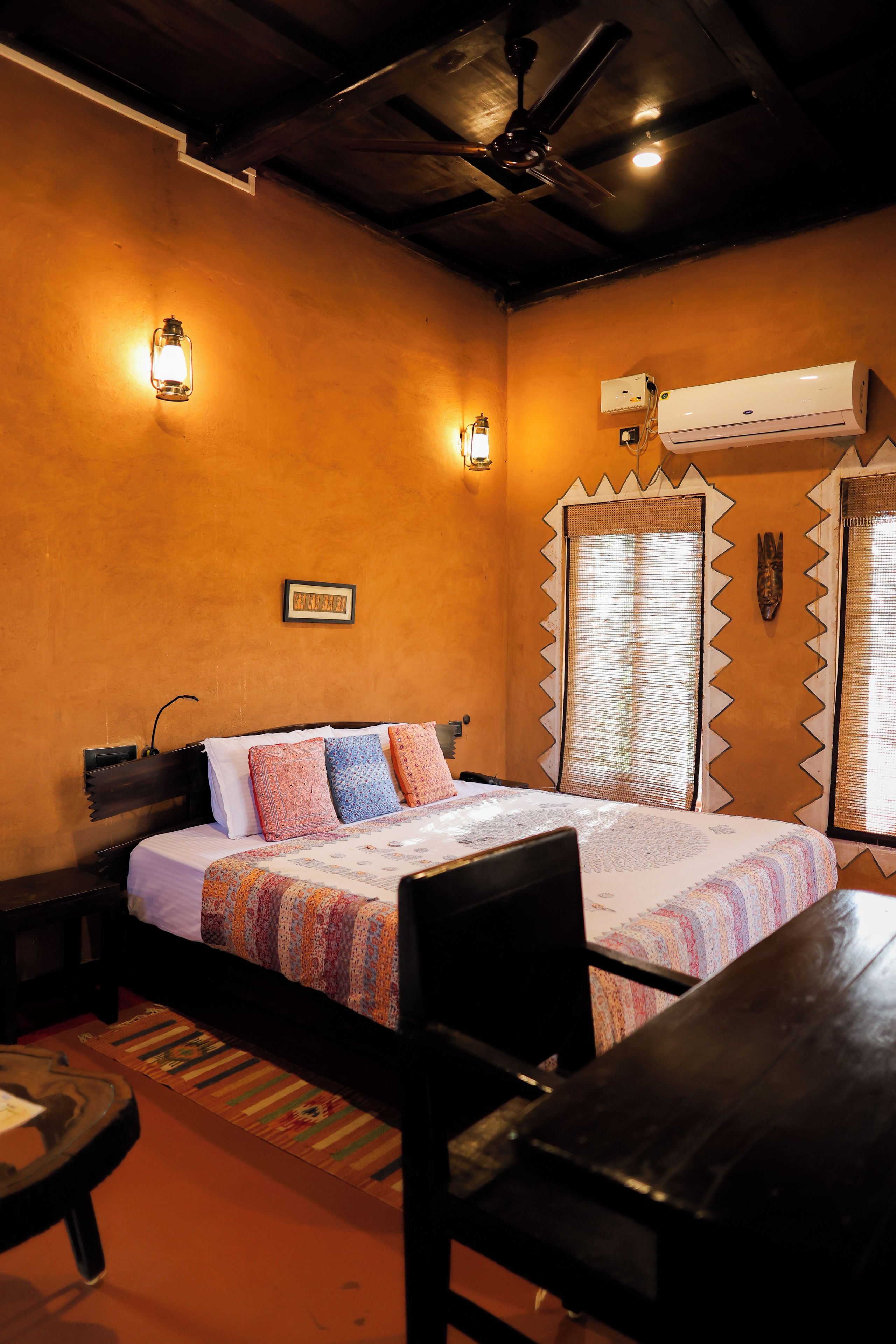 A hotel room with a bed, a table, a chair, a ceiling fan, wall-mounted lights, an air conditioning unit, and a window with a decorative frame.
