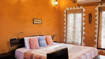 A hotel room with a bed, a table, a chair, a ceiling fan, wall-mounted lights, an air conditioning unit, and a window with a decorative frame.
