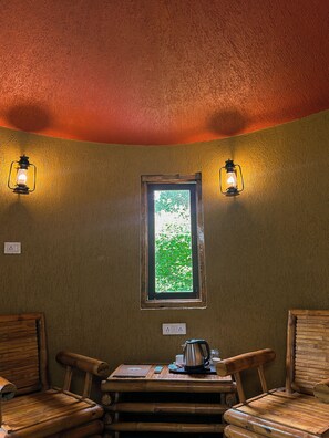 A corner room with a wooden table, two chairs, a window with a view of greenery, and two wall-mounted lights.