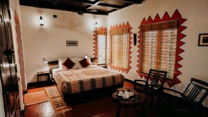 Un dormitorio con cama, dos mesitas de noche, una silla, una pequeña mesa con bandeja y una ventana con cortina estampada.