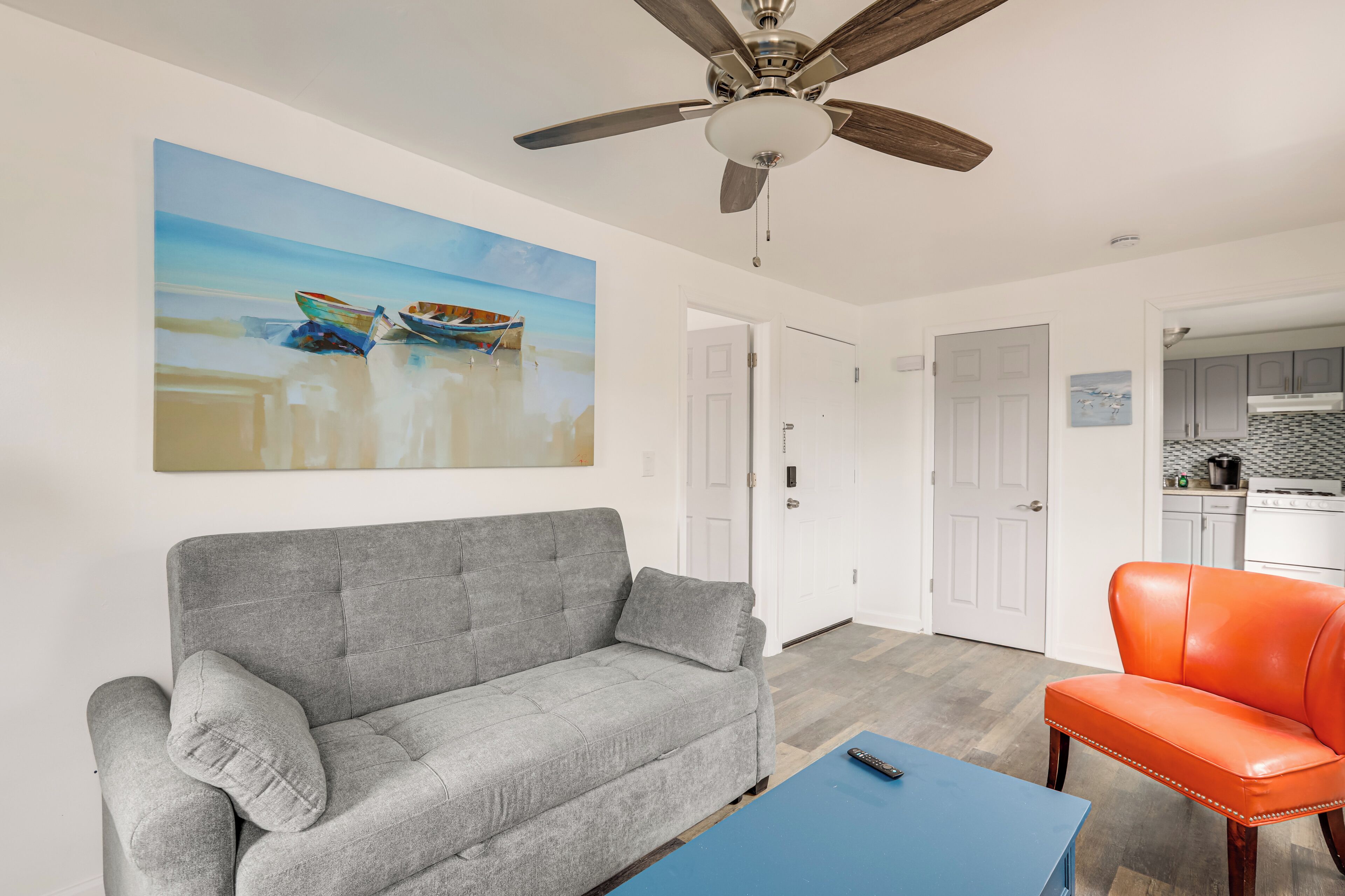 Ein Wohnzimmer mit grauem Sofa, einem orangefarbenen Sessel, Deckenventilator und einem Gemälde mit Booten an der Wand.