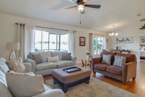 A spacious living room with a ceiling fan, large windows, a brown leather sofa, a grey sofa, and a coffee table. - Work & Play in Apopka! Cozy Extended-stay Home (Apopka)