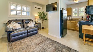 A living room with a leather sofa, a coffee table, a rug, a TV mounted on the wall, and a kitchen area with cabinets and appliances. - 1 Mi to Vanderbilt Beach! Walkable Couples Haven (Naples)