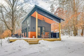 Una cabaña moderna con terraza de madera, ventanas amplias y zona de estar exterior cubierta.