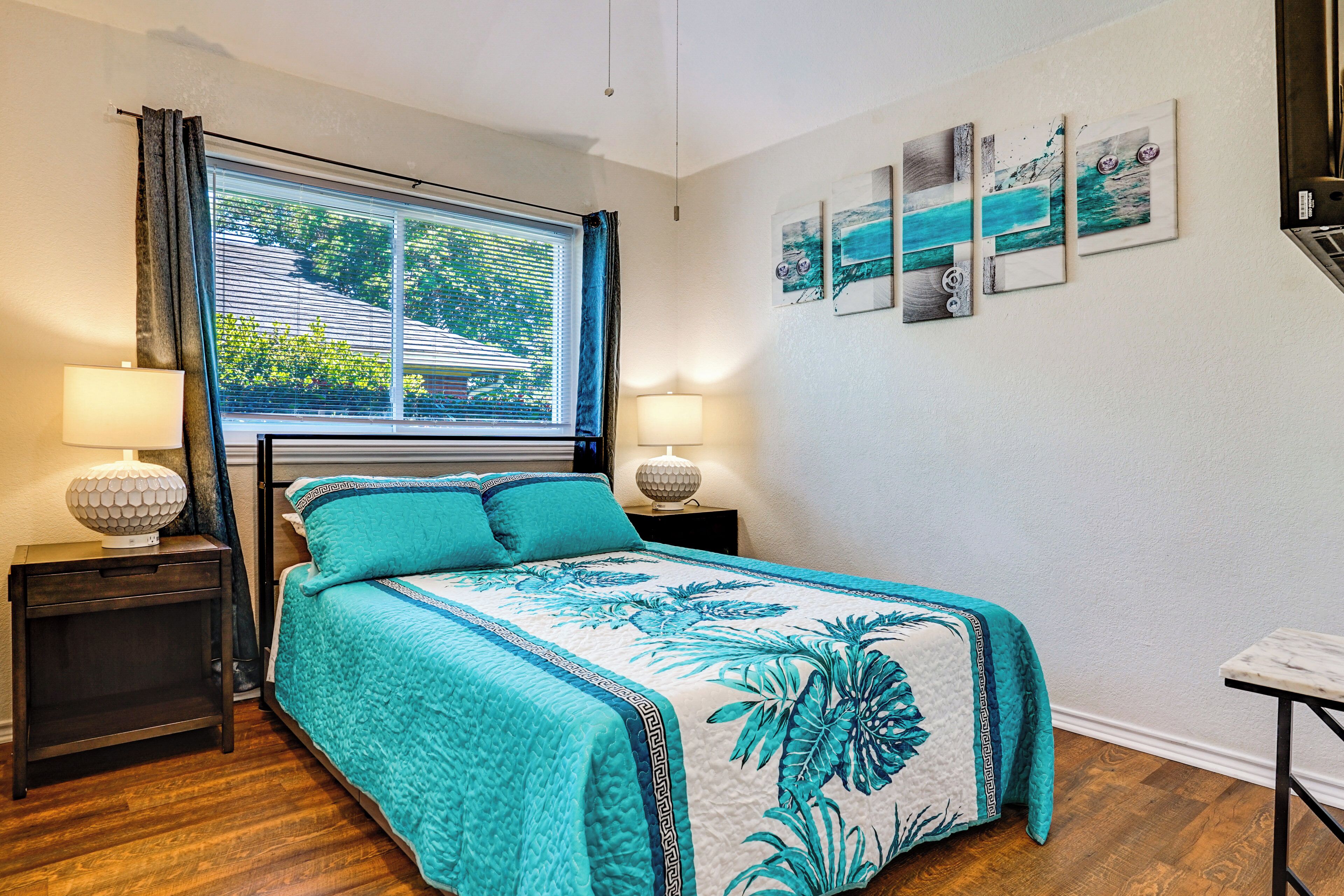 Un dormitorio con una cama, dos mesitas de noche con lámparas, una ventana con cortinas y una obra de arte en la pared.