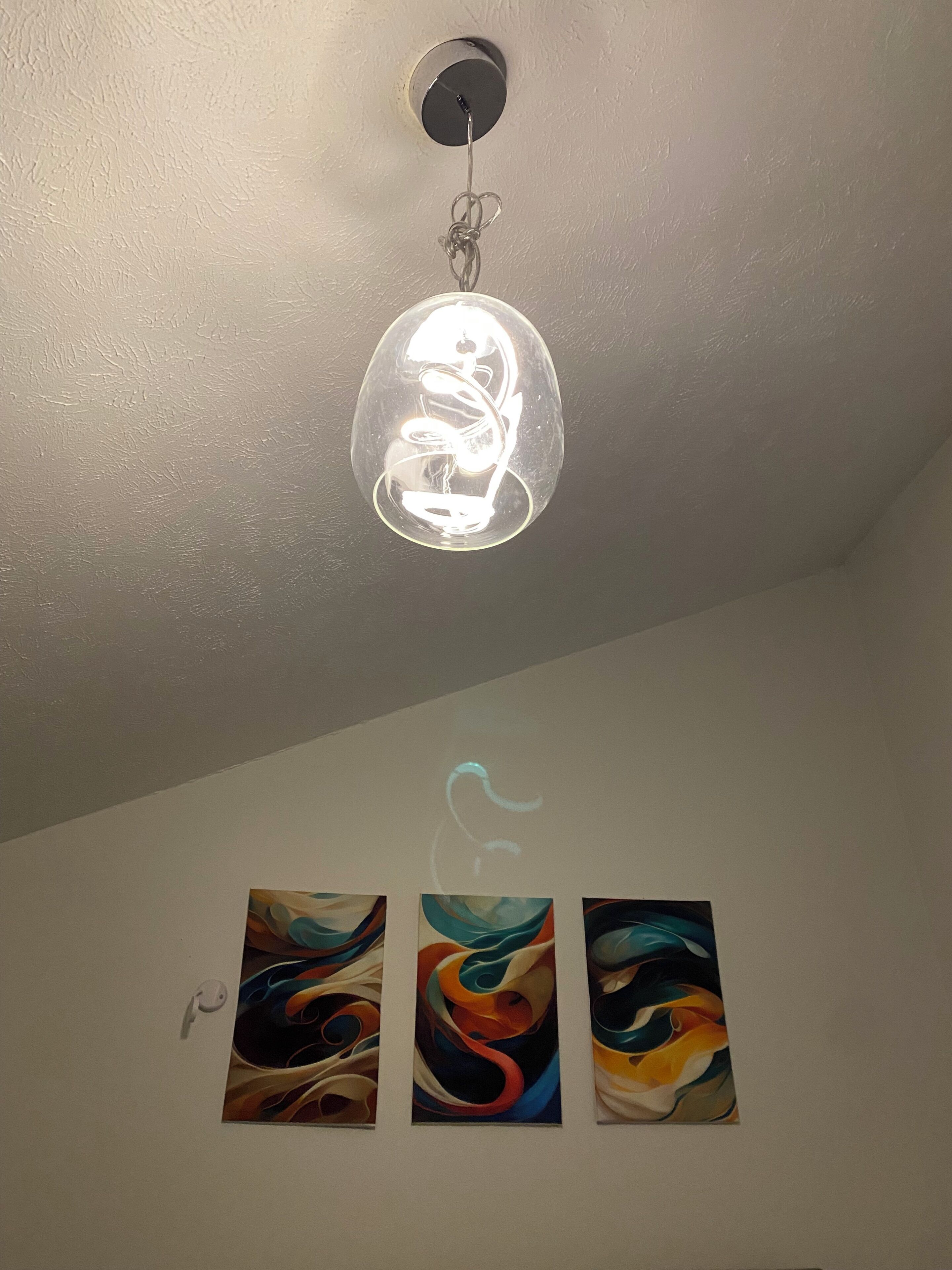 A ceiling light fixture with a clear glass cover, three abstract paintings on a white wall, and a white switch.