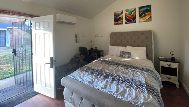 A bedroom with a bed, bedside tables, a chair, a door, and a wall-mounted air conditioner.