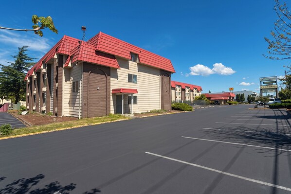 A row of motel buildings with red roofs and beige siding, a parking lot, and a clear blue sky. - Tacoma Port Inn Fife I-5 (Fife)