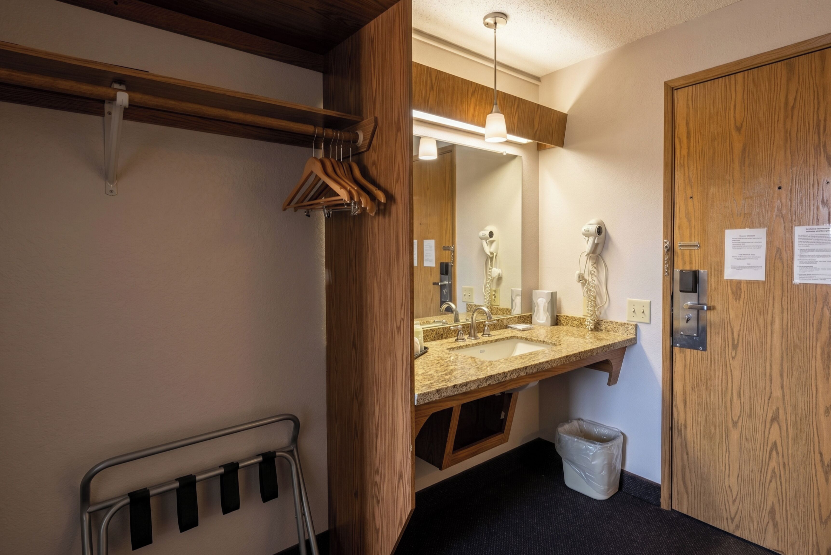 A bathroom with a wooden vanity, a mirror, a sink, a hanging towel rack, a trash bin, and a door with a lock and a paper on it.