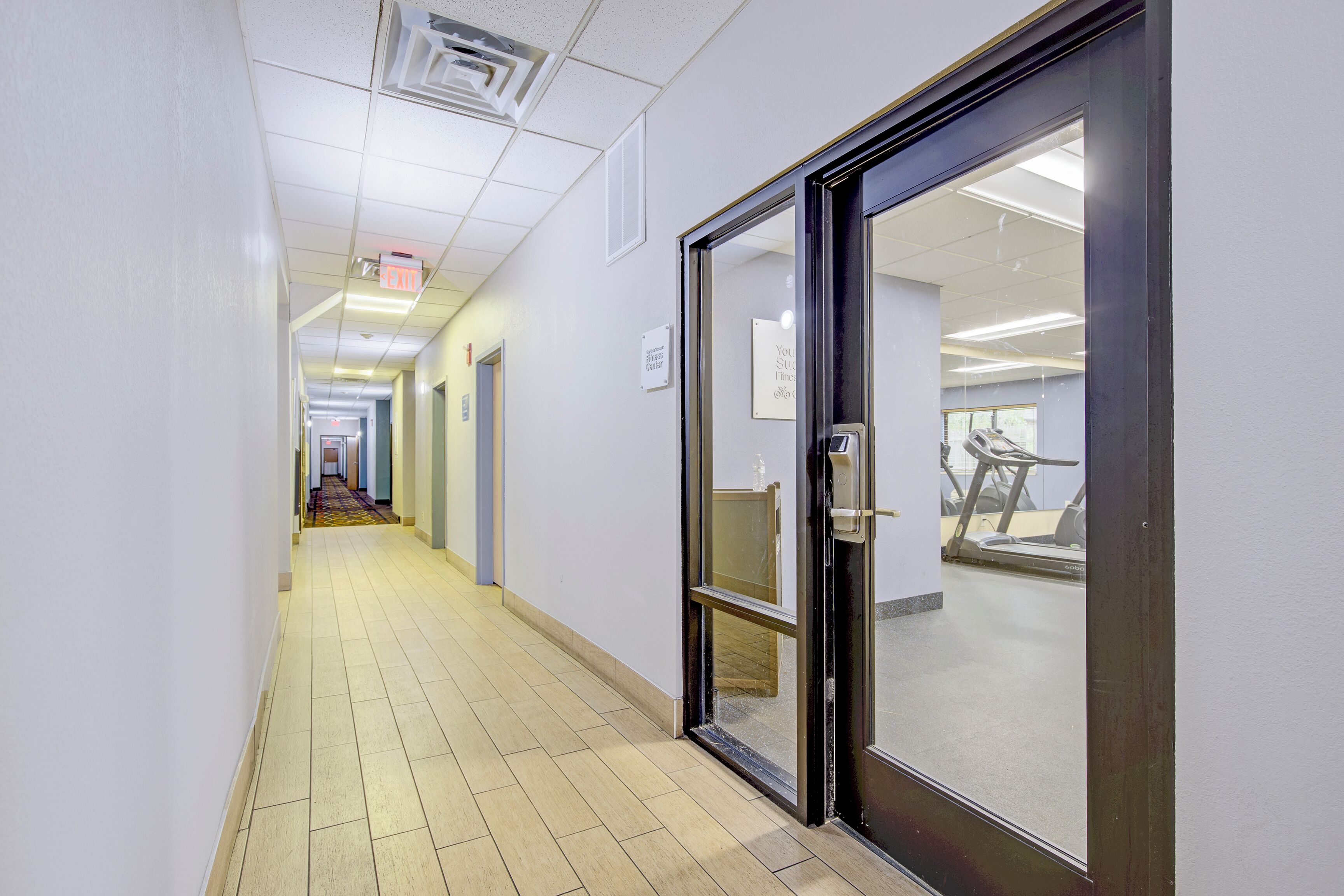 A corridor with a glass door leading to a fitness center, a tiled floor, and a ceiling with fluorescent lighting.