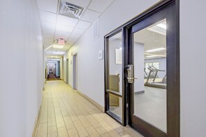 A corridor with a glass door leading to a fitness center, a tiled floor, and a ceiling with fluorescent lighting. - Saint Joseph Inn & Suites Benton Harbor Near I-94 (Benton Harbor)