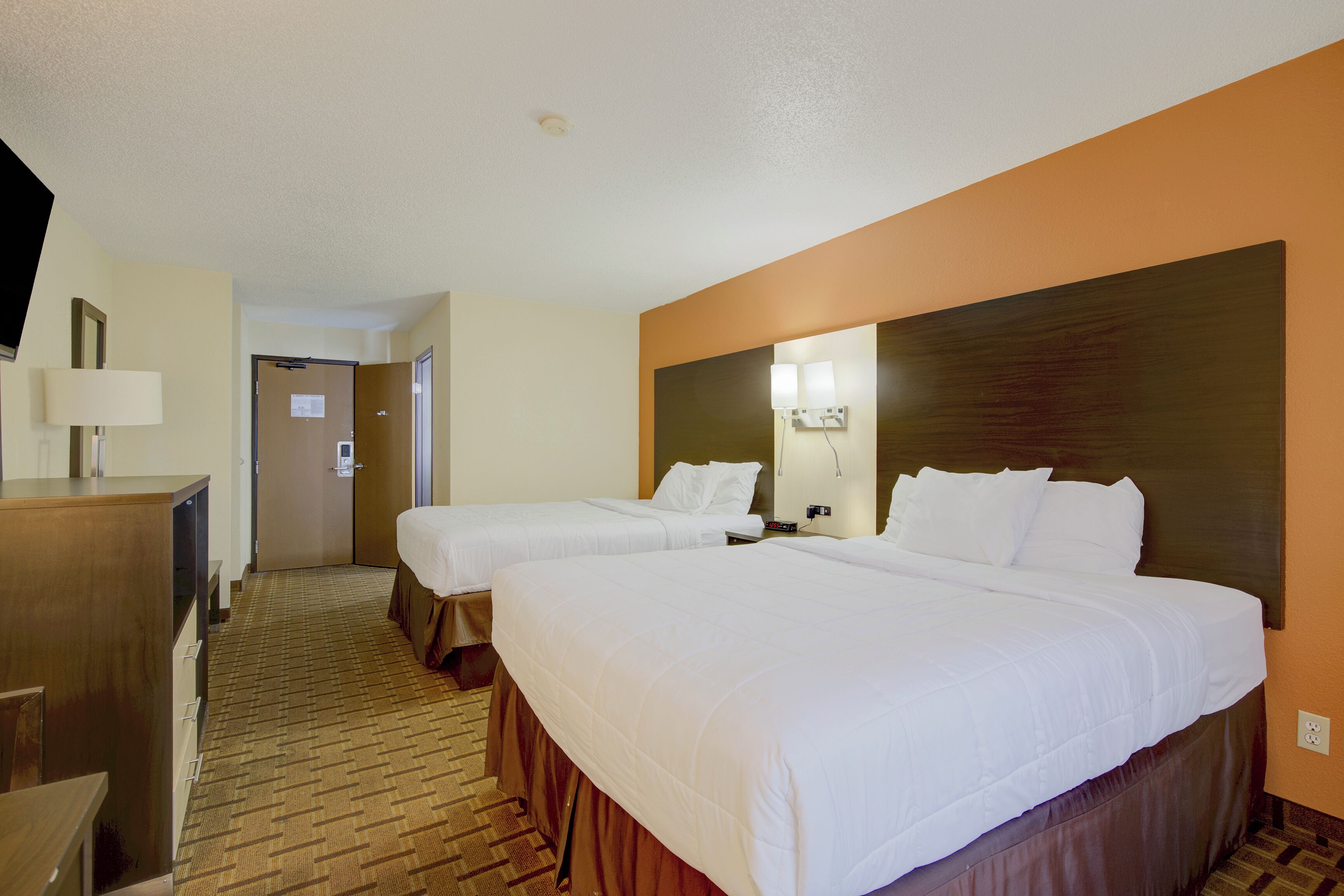 A hotel room with two beds, a desk, a mirror, and a wall-mounted light fixture.