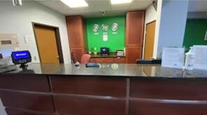 A reception desk with a computer monitor, a green wall with clocks, and a wooden door.