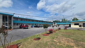 Un motel con fachada azul y blanca, un estacionamiento y un pequeño jardín con arbustos y árboles jóvenes.